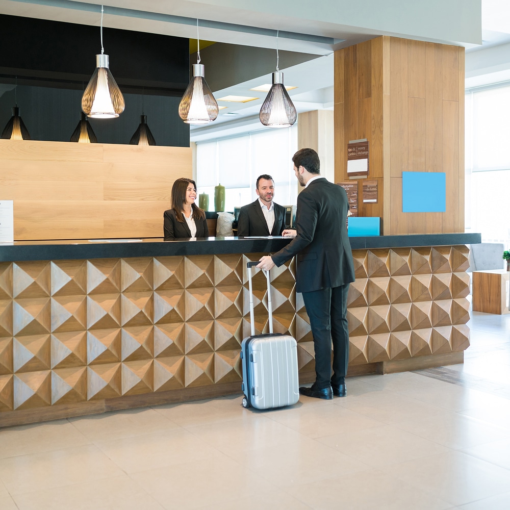 Client avec valise interagit avec deux réceptionnistes d'hôtel souriants à un comptoir d'accueil moderne. Enregistrement professionnel.