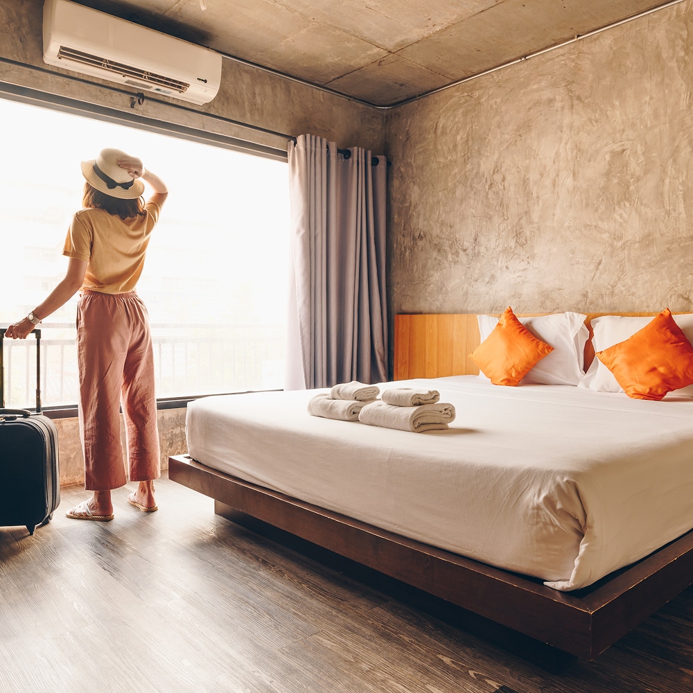 Femme en chapeau regardant par la fenêtre dans une chambre d'hôtel moderne avec lit et valise.
