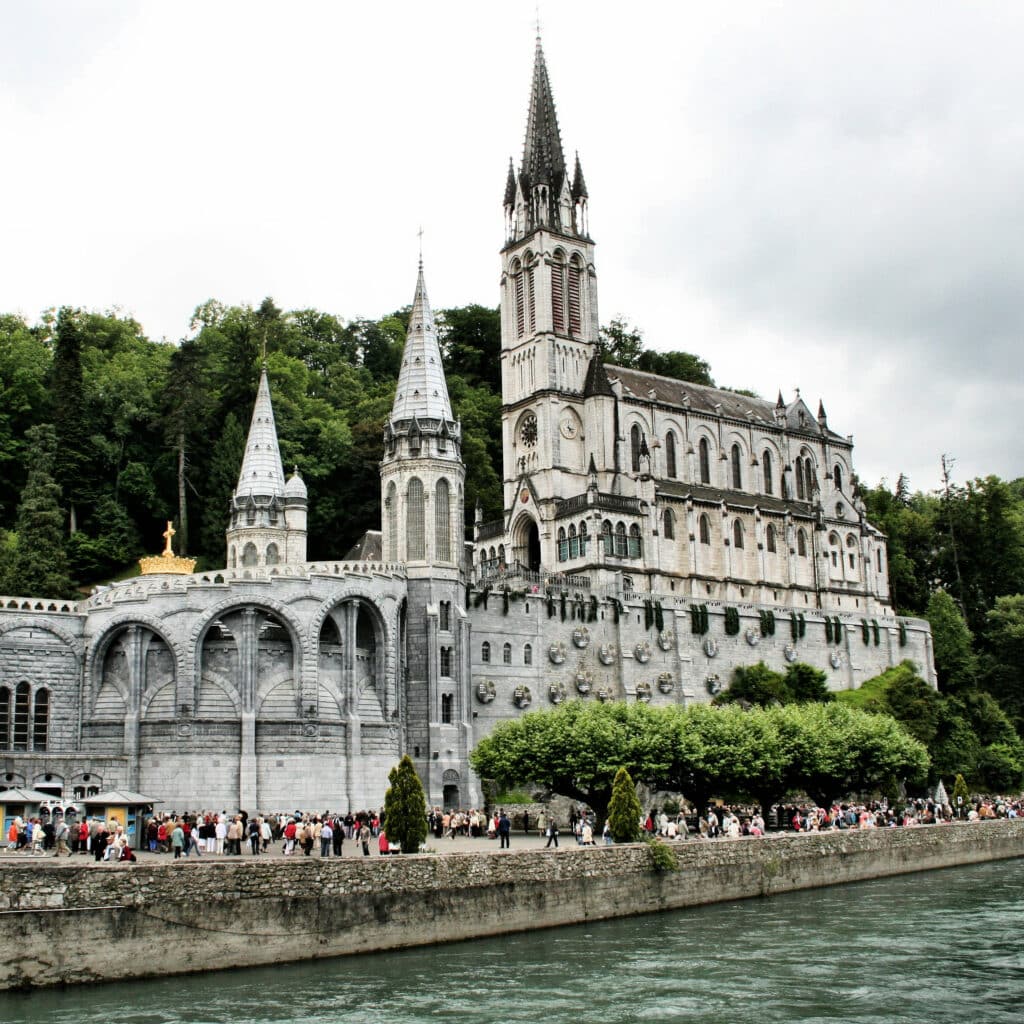 Grande basilique gothique grise du Sanctuaire de Lourdes avec tours pointues, surplombant une rivière et des pèlerins.