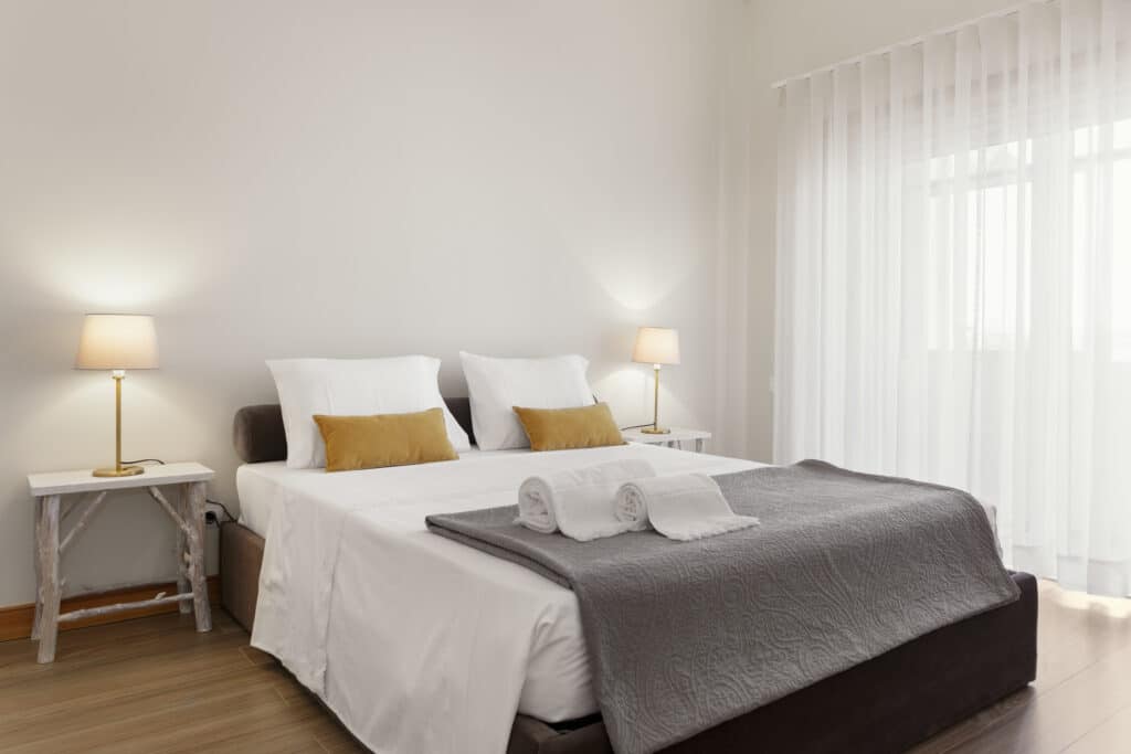 Chambre moderne avec lit double blanc, coussins moutarde, jeté gris texturé. Tables de chevet bois clair, lampes chaudes, rideaux blancs.