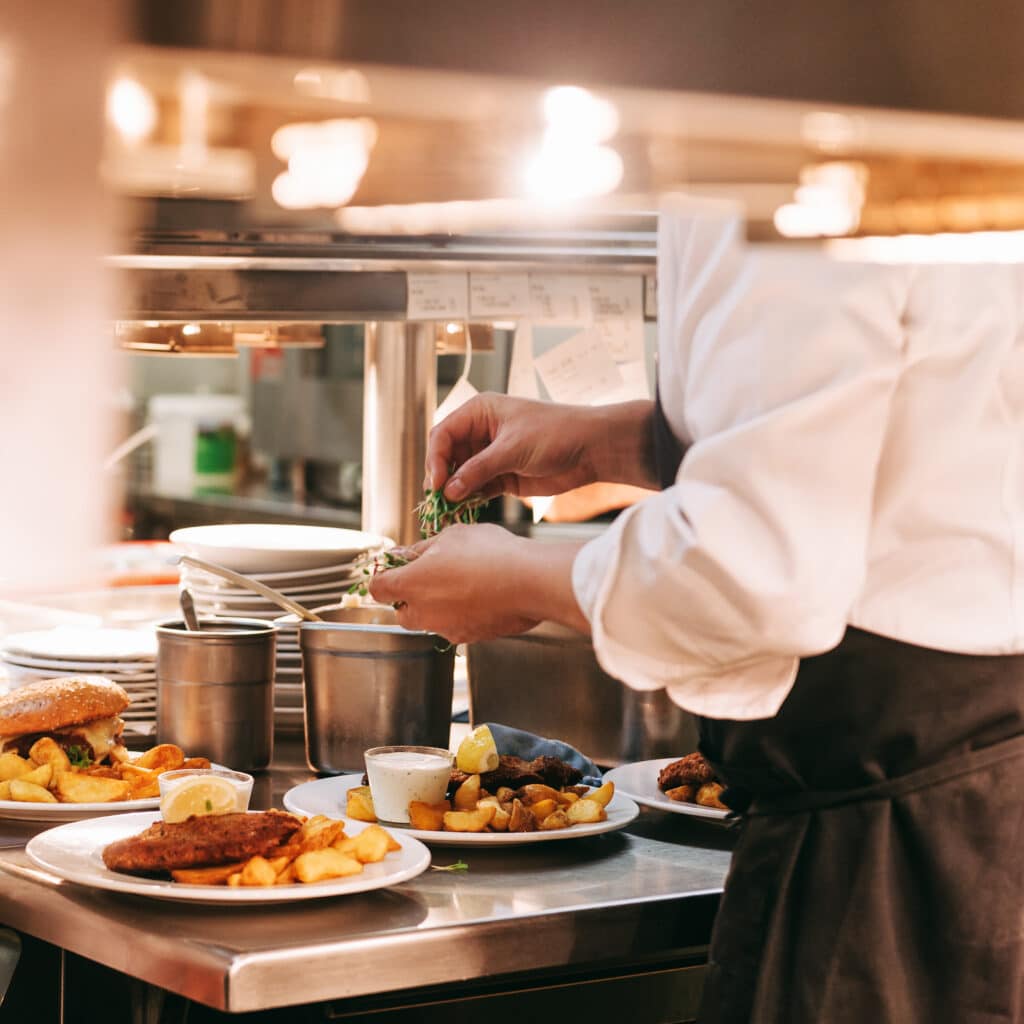 Chef garnissant un plat de schnitzel et frites avec des herbes en cuisine professionnelle. Un burger est aussi sur le comptoir.