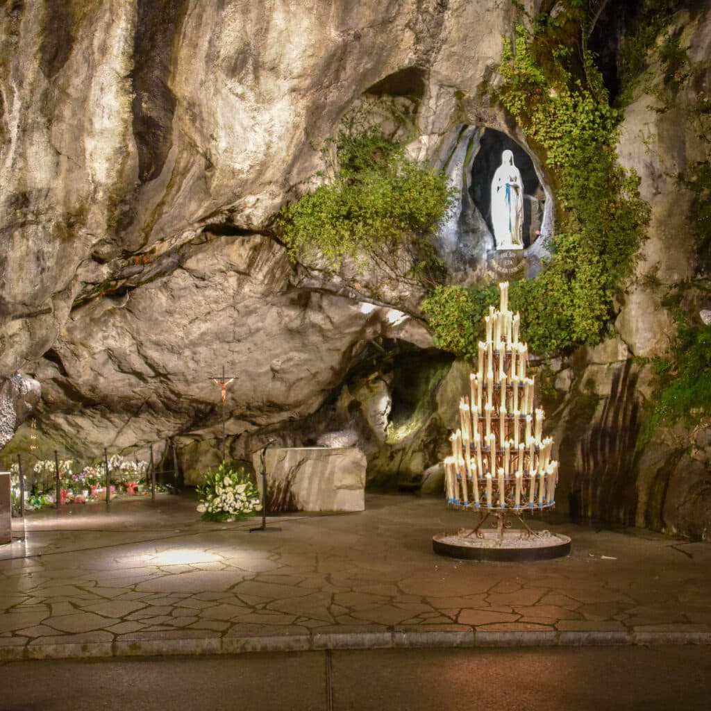 Grotte de Lourdes avec statue de la Vierge Marie, inscription "QUE SOY ERA IMMACULADA COUNCEPCION", un grand chandelier allumé et autel. Atmosphère sacrée.