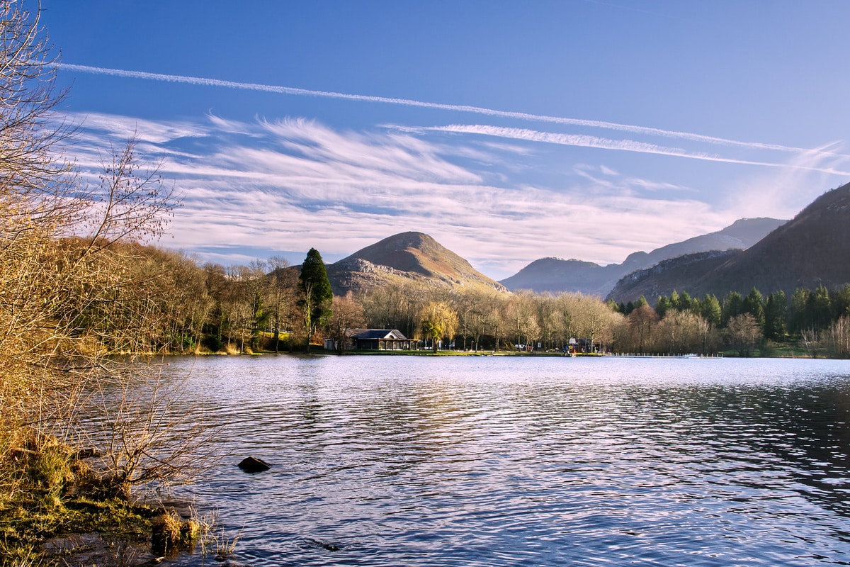Lac paisible entouré de montagnes et d'arbres dépouillés en hiver. Le ciel bleu est parsemé de nuages et de traînées d'avion.