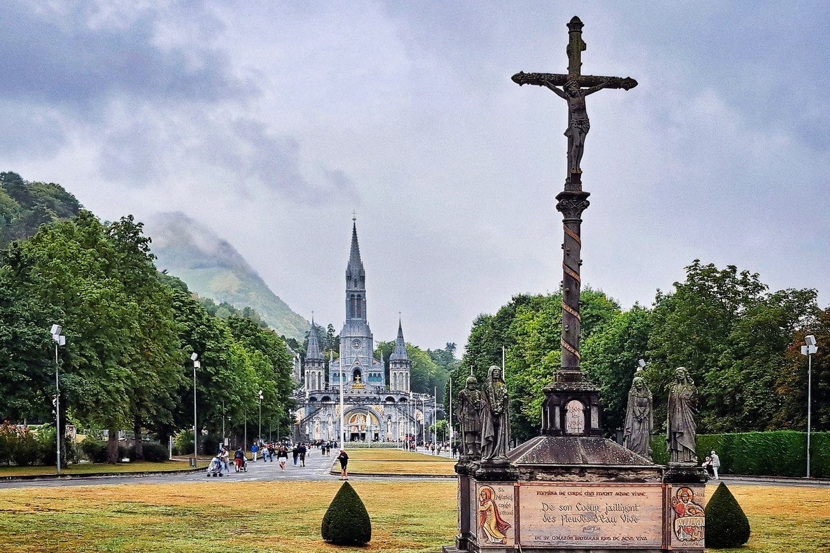 Grande croix en pierre avec Christ, devant la Basilique du Rosaire et la foule à Lourdes. Inscription 'De son Coeur jailliront des Fleuves d'eau Vive'.