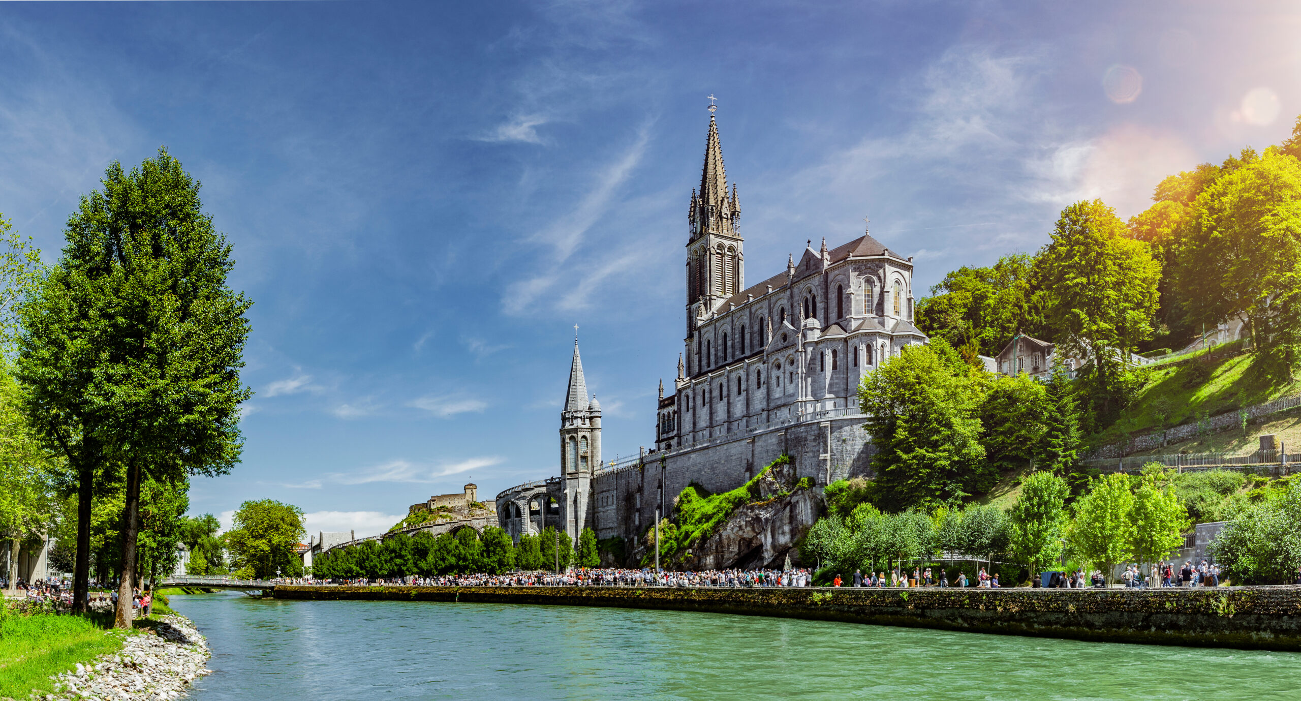 Vue panoramique du Sanctuaire de Lourdes avec la basilique majestueuse et des pèlerins marchant le long de la rivière Gave de Pau par temps ensoleillé.