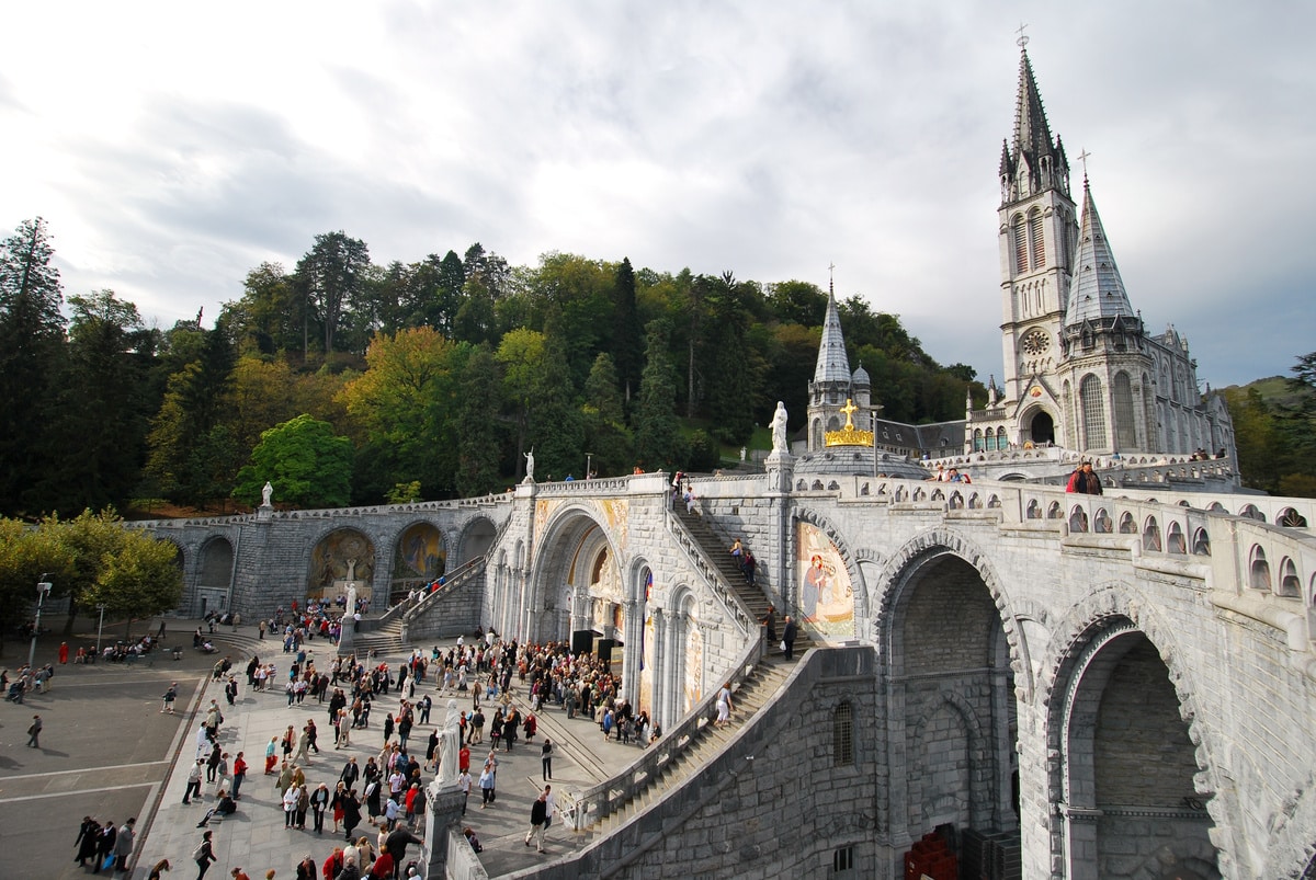 Grande basilique de Lourdes en pierre, avec ses spires et arches, surplombant une esplanade animée par de nombreux pèlerins.