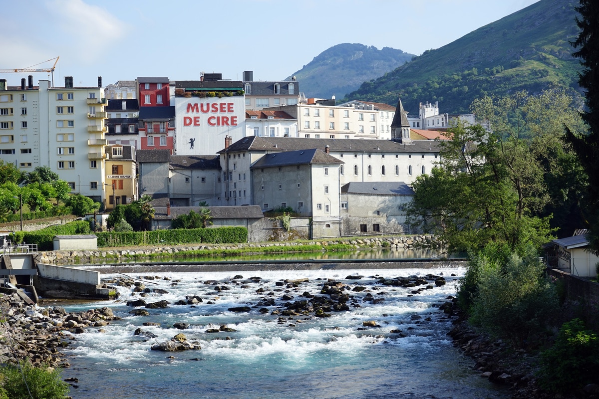 Rivière turquoise tumultueuse et rochers, avec des bâtiments historiques en arrière-plan, dont le 'Musée de Cire' et ses montagnes.