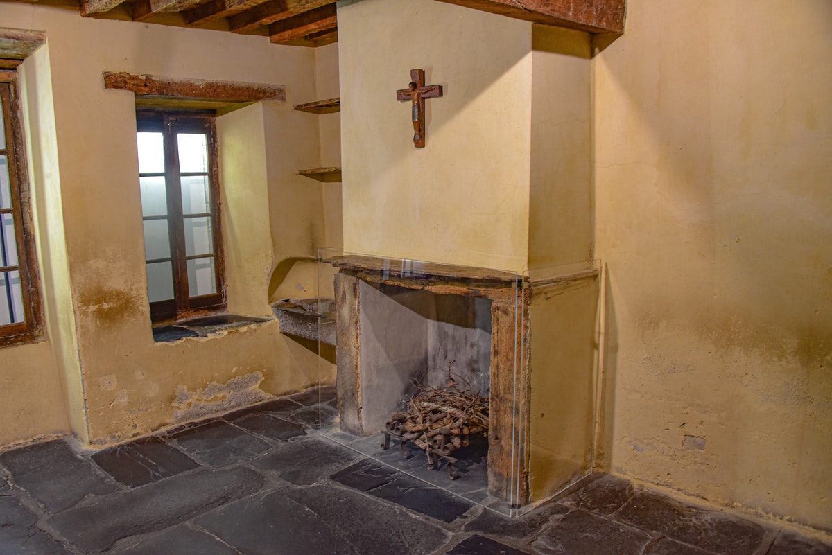 Pièce ancienne rustique avec murs jaunes usés, sol en pierre. Cheminée avec bûches derrière verre, crucifix mural. Fenêtres en bois.